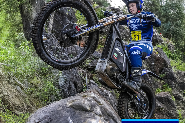Motorcyclist wearing blue gear riding over rocks in a forest setting.