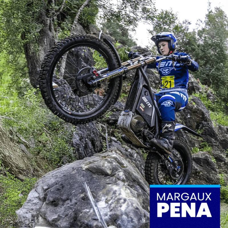 Motorcyclist wearing blue gear riding over rocks in a forest setting.