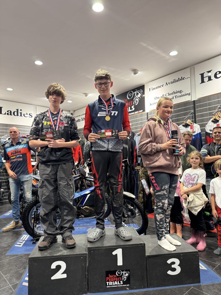 Three young people stand on a podium holding certificates, wearing medals, in a motorcycle store setting.