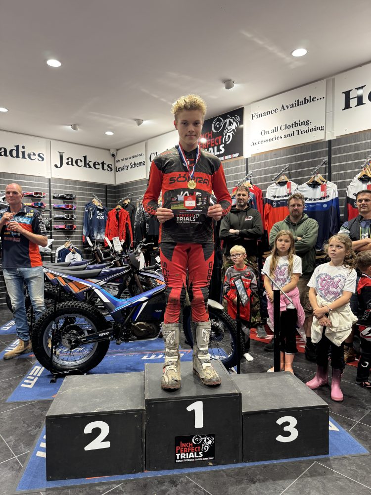 Person on podium in motocross gear holding trophy, surrounded by people indoors.