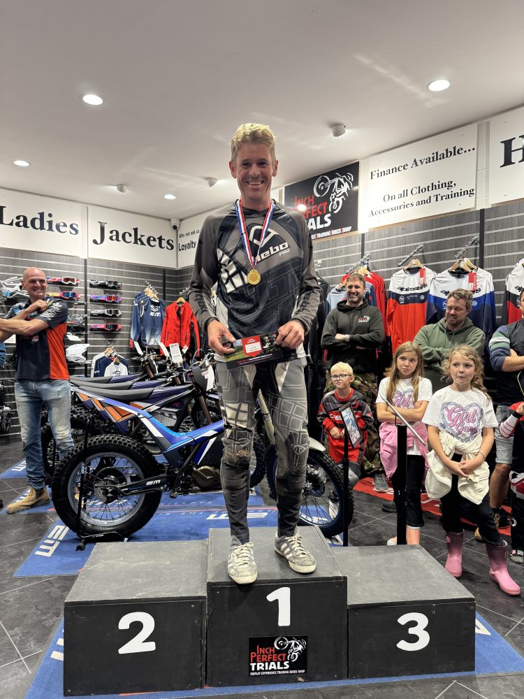 Person on podium holding medal, wearing racing gear, surrounded by onlookers in a store with motorbikes.