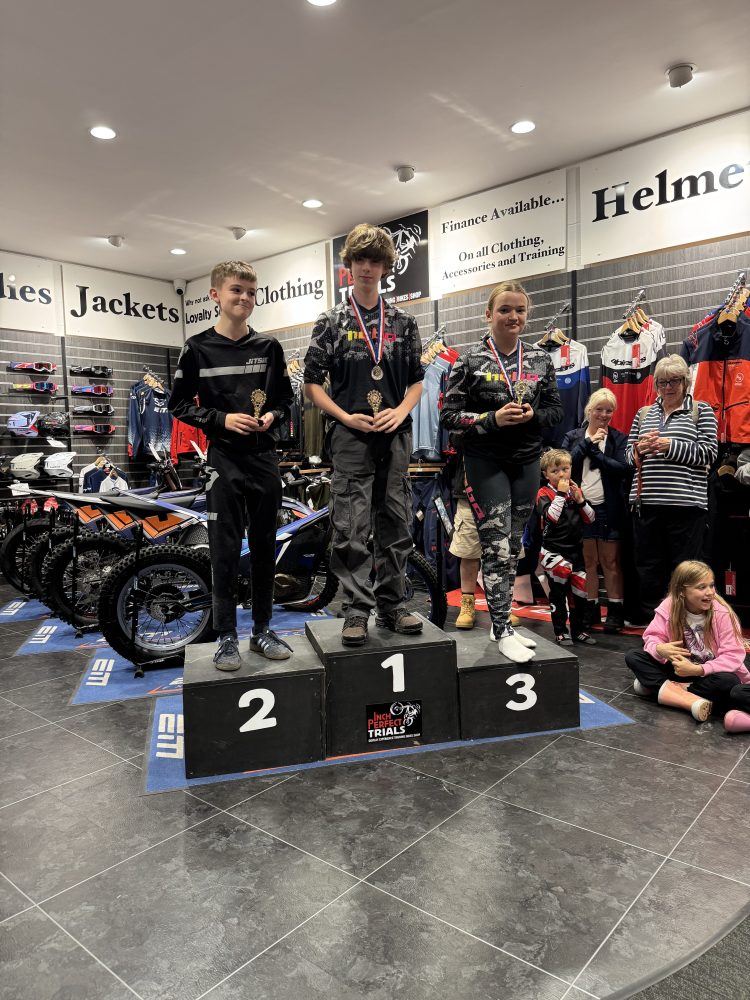 Three young winners with medals on podium in motorcycle gear inside a bike shop.
