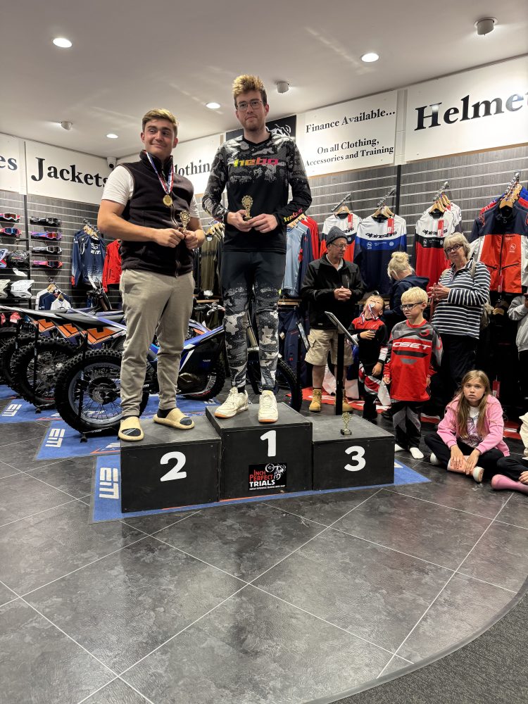 Two people on a podium holding trophies in a motorcycle gear store with an audience.