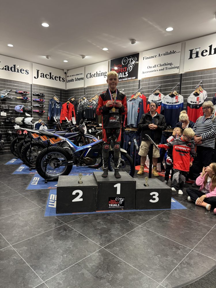 A child stands on a podium holding a medal in a motorcycle shop with spectators and bikes in the background.