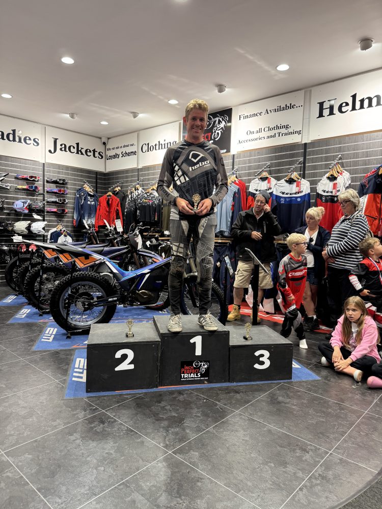 Man in racing gear on podium with bikes and people in store backdrop.