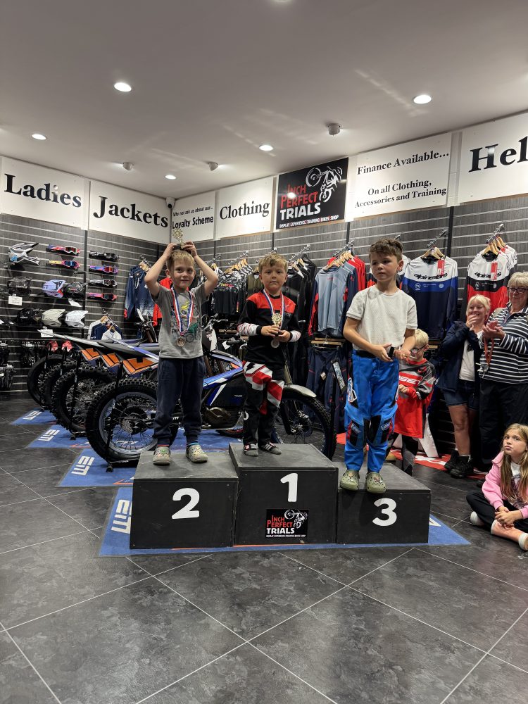 Three kids on a winners' podium in a motorcycle shop, holding medals. Background shows clothing and equipment.