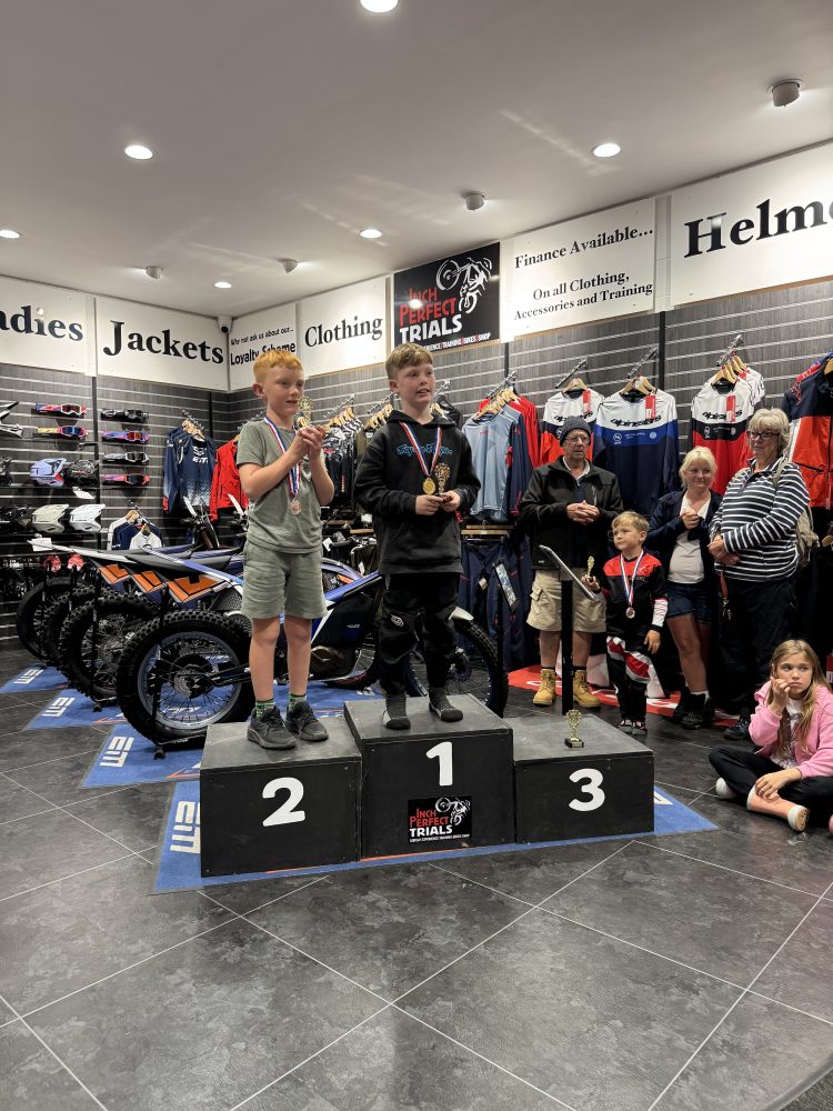 Two boys on a podium with medals in a motorcycle store; others watch nearby.