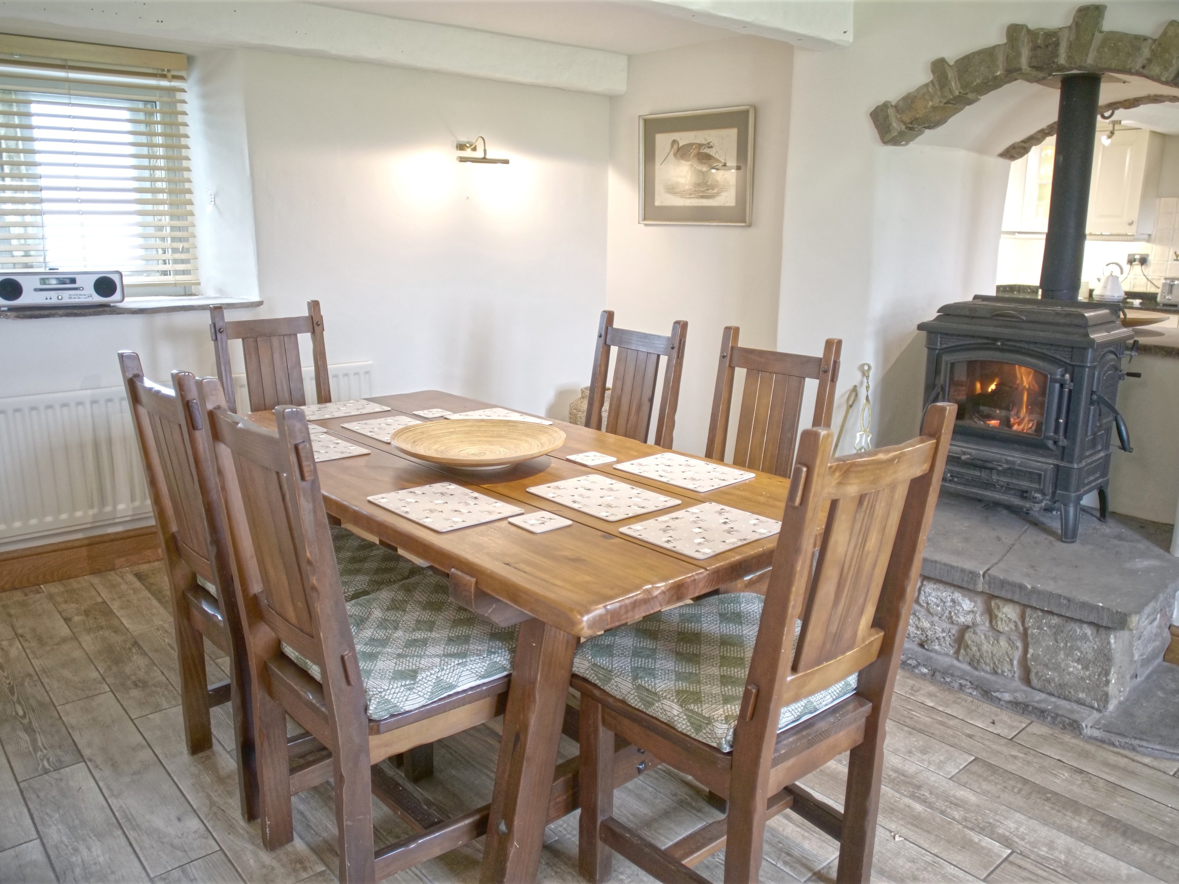 Cozy dining area with wooden table, six chairs, wood burner, and window with blinds.