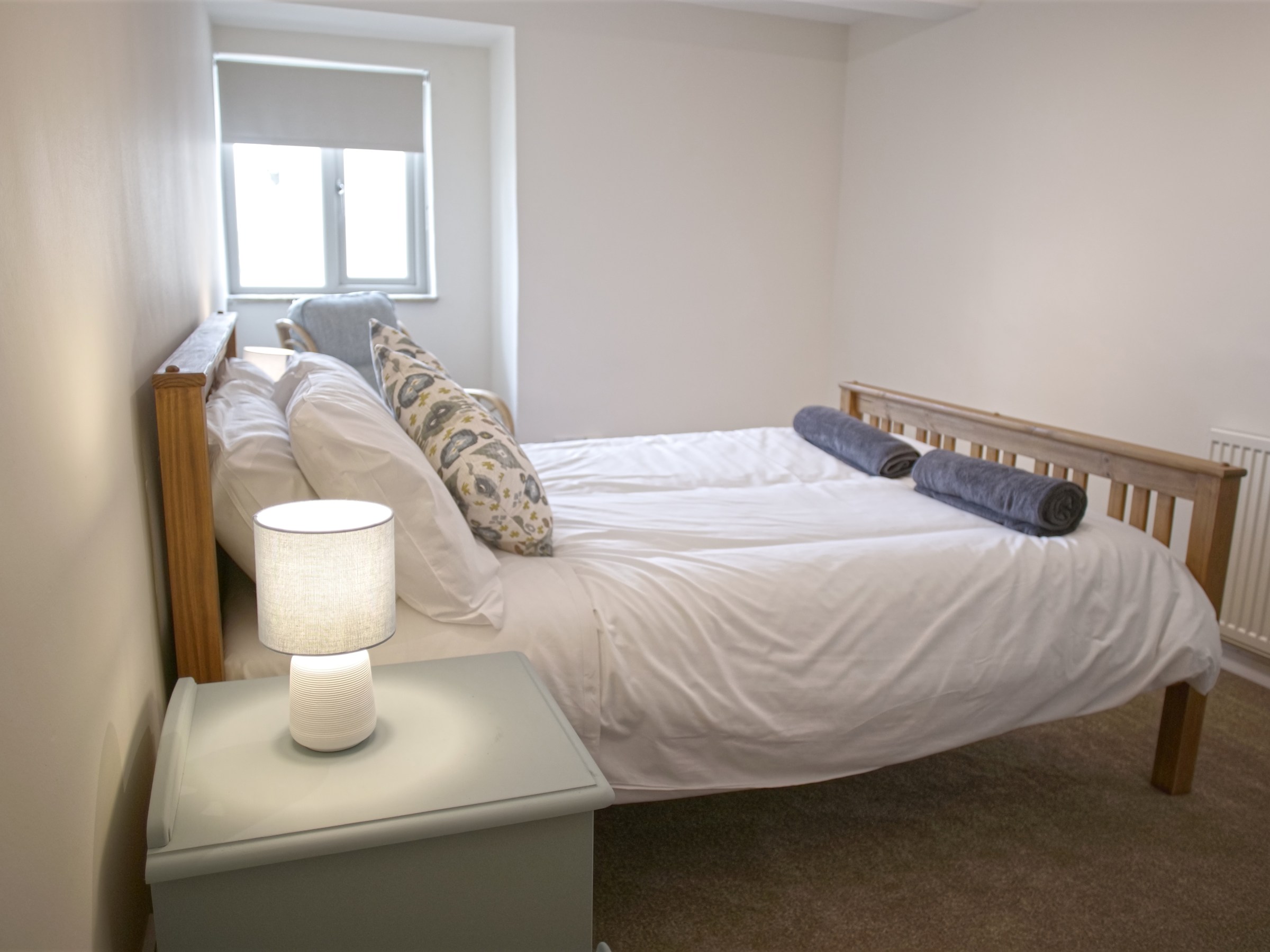 Small bedroom with a wooden bed, side table with lamp, and window with blind.