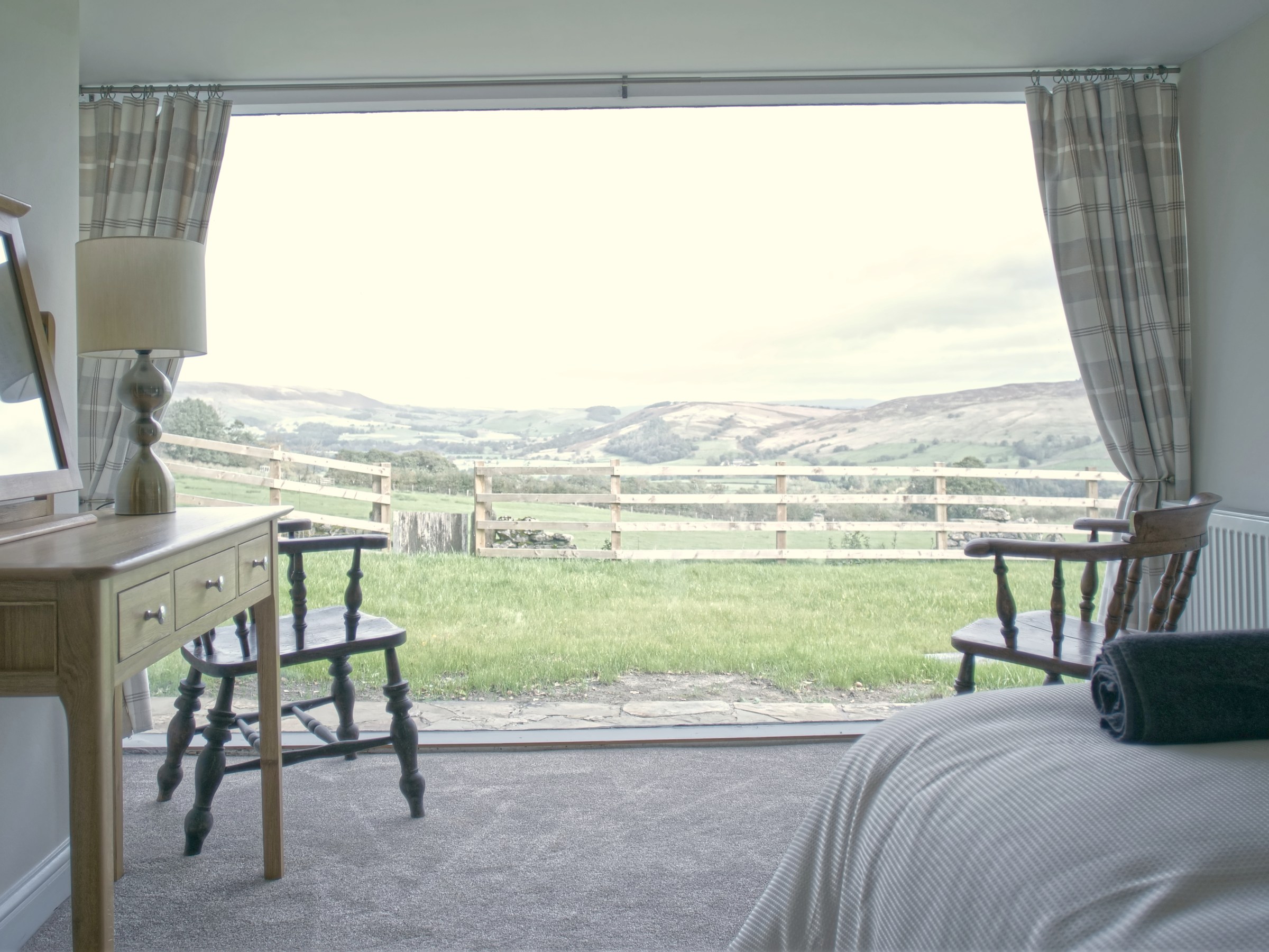Bedroom with large window view of hills, two chairs, a table, and a bed with folded towel.