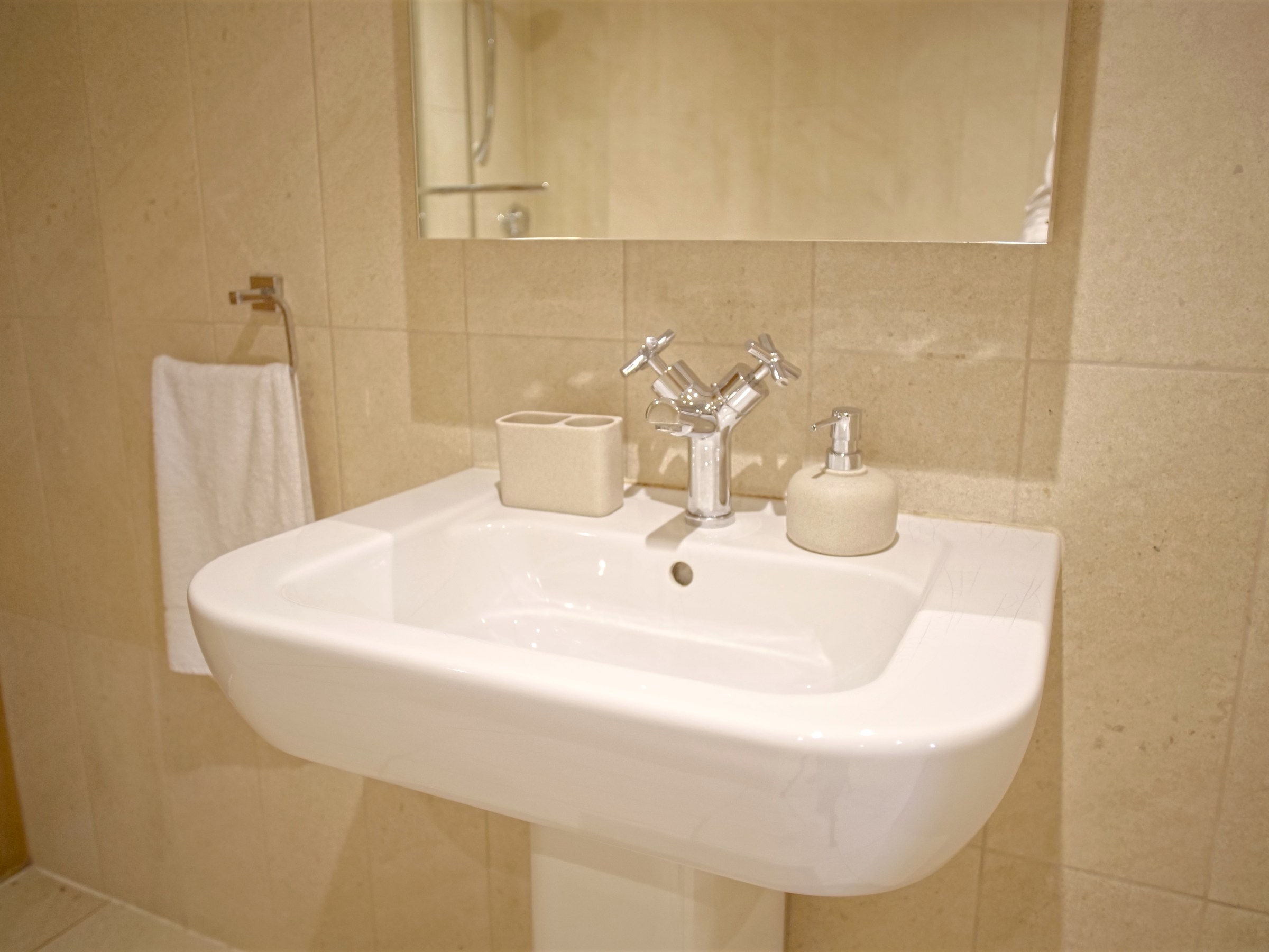 Bathroom sink with towel, soap dispenser, and mirror on beige tiled wall.