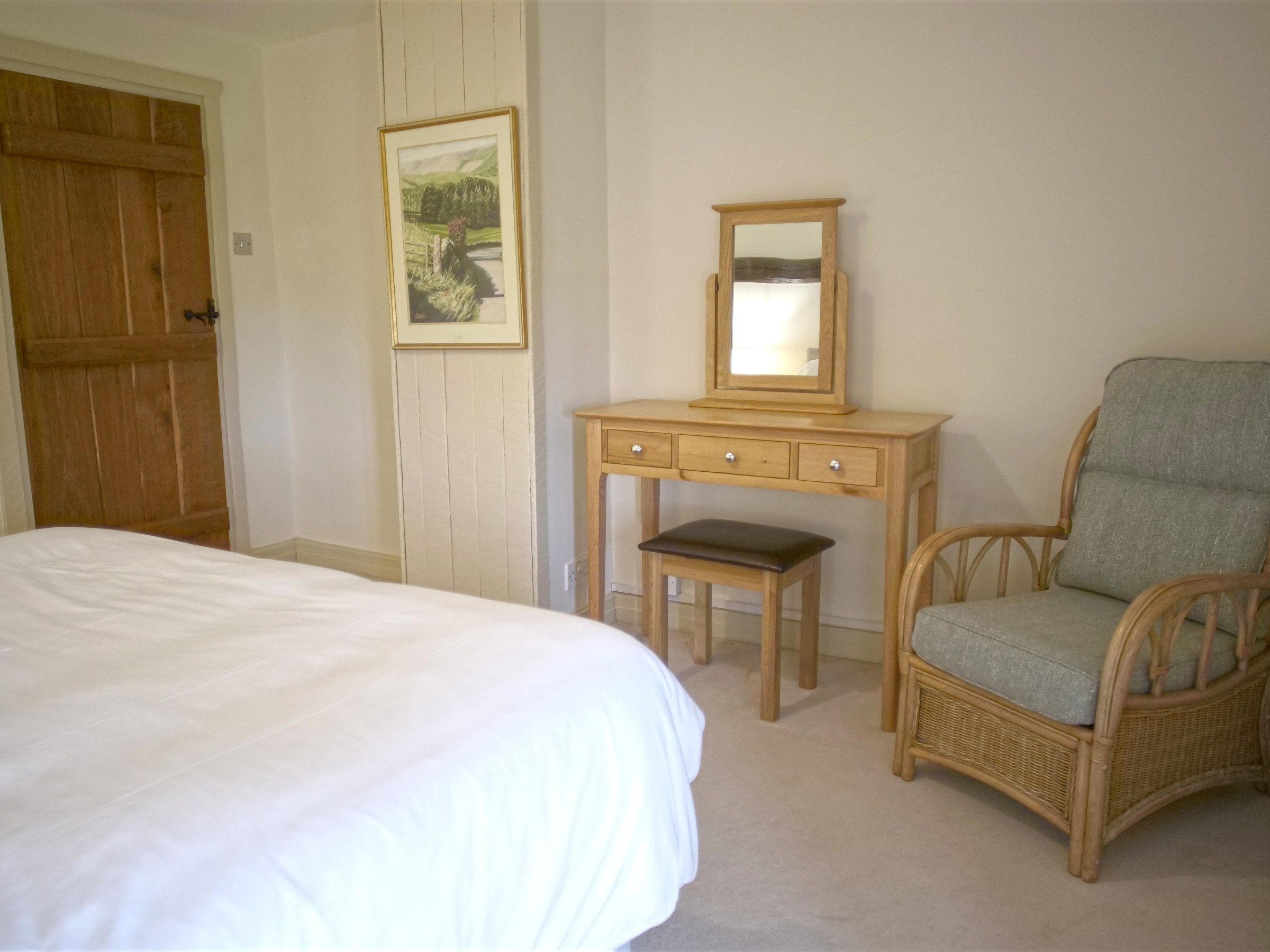 Bedroom with a bed, wooden door, framed picture, dresser with mirror, stool, and wicker chair.